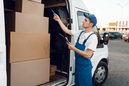 送货员在车内的制服检查箱，送货服务。男子站在纸板包裹在车辆，男性送货，信使或运输工作