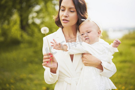 绿色春光摄影照片_年轻漂亮的母亲和可爱的小女儿在绿色的草地上吹向蒲公英的户外时尚肖像。春光映象
