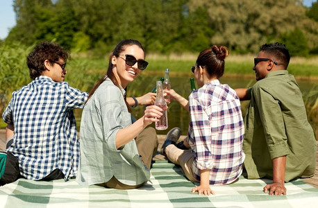 苹果酒摄影照片_休闲、野餐和人的概念-朋友们在夏季公园的湖畔码头喝啤酒和苹果酒。朋友们在湖边码头喝着啤酒和苹果酒