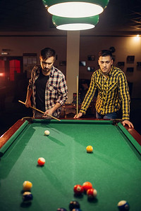 游戏室摄影照片_两个男性台球运动员与线索在桌子与彩色球，台球室。男子在体育酒吧玩美国台球游戏