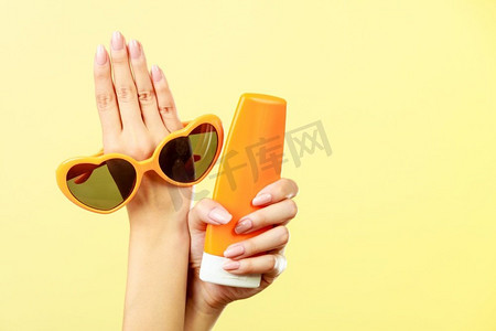假日夏日时尚护眼护肤理念。手持心形太阳镜的女子防晒霜，黄色背景