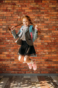 可爱的女学生与书包跳在砖墙。顽皮的女学生与背包姿势在学校