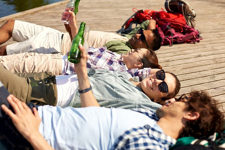 休闲、野餐和人的概念-朋友们在夏季公园的湖畔码头喝啤酒和苹果酒。朋友们在湖边码头喝着啤酒和苹果酒