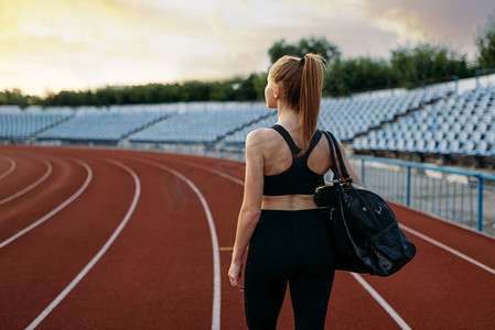 女跑步者在运动服持有运动袋，背面视图，在体育场训练。妇女做伸展运动之前运行在户外竞技场