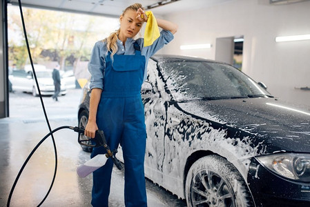 疲惫的女洗衣工穿着制服，手里拿着泡沫枪，洗车。女人洗车、洗车站、洗车生意