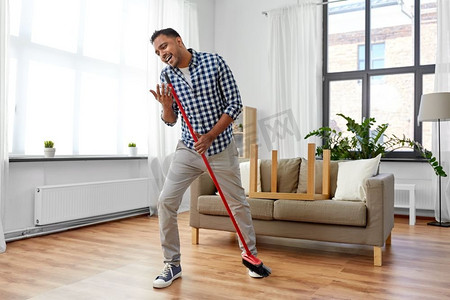 清洁、家务和家政概念—印度男子用扫帚扫地和唱歌在家里。男人用扫帚打扫和唱歌在家里
