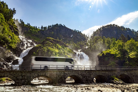 旅游巴士摄影照片_旅游巴士行驶在公路Latefossen瀑布Odda挪威。Latefoss是一个强大的双瀑布。