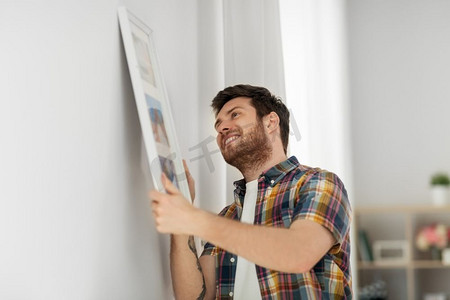 家里挂画摄影照片_室内装饰和装修概念—微笑的男子挂在框架的图片到墙上在家里。一个人在家里把画挂在相框里的墙上