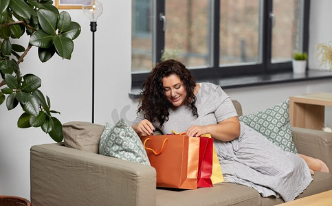 消费主义，人和销售概念—快乐的微笑的年轻妇女与购物袋在沙发上在家里。快乐的年轻女人与购物袋在家里