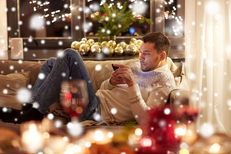 圣诞节、科技和节日--雪天在家用智能手机发短信的概念人。男人带着智能手机在家里过圣诞节