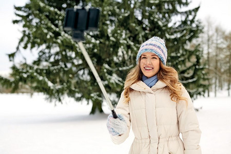 人、季节、休闲理念--冬天快乐微笑的女人拿着自拍杆拍照。冬天，一名女子用自拍杆拍照