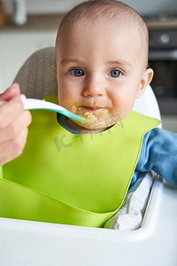 微笑的婴儿男孩的肖像在家里高椅子被喂固体食物由母亲用勺子  