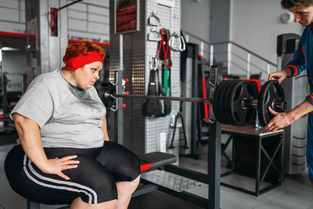 超重出汗的妇女在健身房与杠铃锻炼。燃烧卡路里，肥胖的女性在体育俱乐部