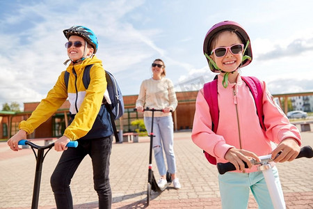 教育，学校和家庭概念—快乐的女儿，儿子和母亲骑滑板车户外。快乐的学生与母亲骑滑板车