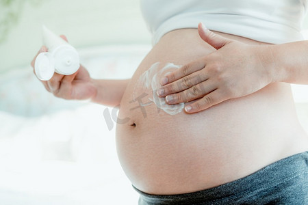 婴儿护肤摄影照片_快乐的孕妇在孕妇肚子上涂抹护肤霜，防止妊娠纹。健康肌肤按摩护理及保湿护理理念。