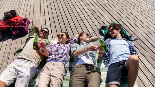 旅行，旅游和人的概念—朋友喝啤酒和苹果酒在木露台在夏天。朋友喝啤酒和苹果酒在木露台