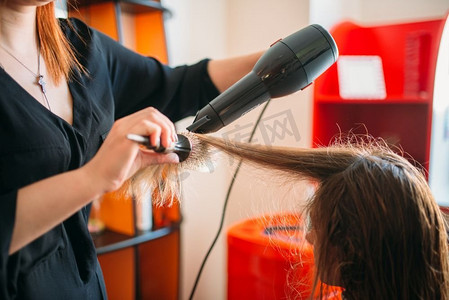 发型师吹头发摄影照片_发型师用吹风机吹干头发，美发沙龙里的女理发师。美容工作室的发型制作