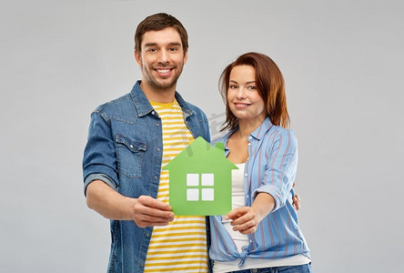 房地产，家庭和生态概念—微笑的夫妇拿着绿色纸房子在灰色背景。微笑的夫妇抱着绿色的纸房子