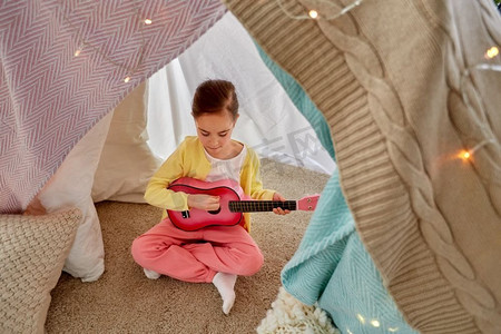 童年和hygge概念—小女孩玩吉他在孩子帐篷或帐篷在家里。小女孩玩吉他在孩子们帐篷在家里