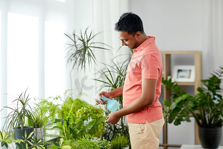 人，自然和植物护理概念—微笑的印度男子喷洒室内植物由喷水器在家里。男子在家中喷洒室内植物