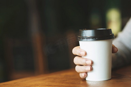 咖啡热咖啡摄影照片_年轻女子手持纸杯的外卖，在咖啡厅的桌子木头上喝着热咖啡。