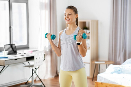 少女肌肌摄影照片_健身，运动和健康的生活方式概念—快乐的少女哑铃在家里锻炼。十几岁的女孩与哑铃锻炼在家里