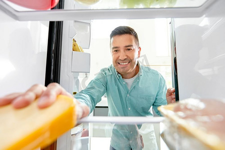 西班牙奶酪摄影照片_食物、饮食和饮食概念—微笑的中年男子从厨房的冰箱拿奶酪。男人从厨房的冰箱里拿食物