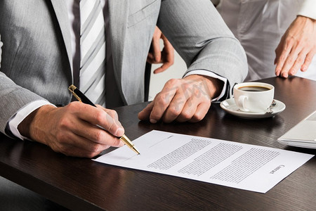 一位穿着灰色西装的商人坐在办公桌前，近距离地签着一份合同。签署合同的商人