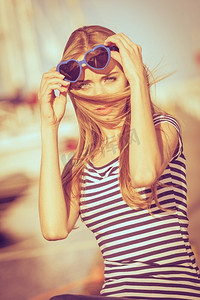 心形太阳镜摄影照片_夏日时尚。戴着蓝色心形太阳镜的肖像女孩在码头的夕阳下享受夏日的微风