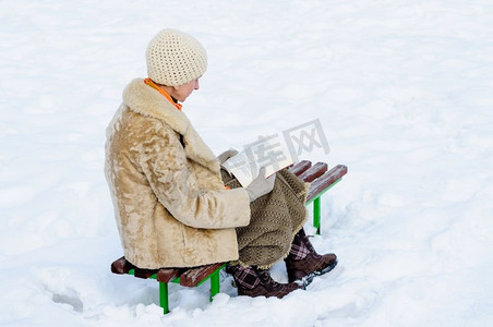 女人坐在长凳上看书在冬天。雪