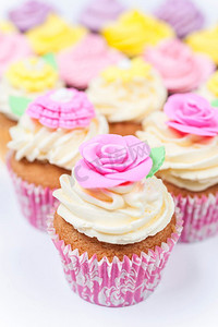 杯蛋糕与糖霜或糖霜，粉红色，紫色，黄色和奶油与绿叶，玫瑰和花卉装饰拍摄在白色背景