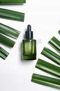 绿色玻璃化妆瓶，白色背景，绿色棕榈叶。使用保湿面部精华液进行皮肤护理。带有复制空间的俯视图。