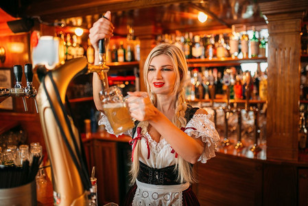 性感的女服务员倒啤酒到杯子在柜台在酒吧。在传统风格的衣服中有吸引力的形状的Fasheberfest酒吧女招待