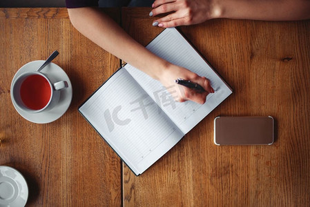 咖啡厅女孩摄影照片_女性手在木桌上的笔记本上写字的俯视图。