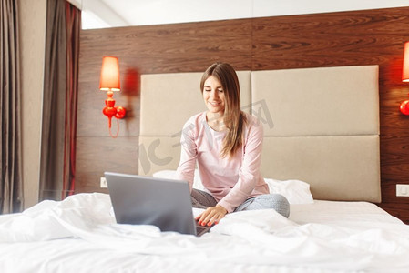 微笑的女人坐在床上，用笔记本电脑，早上好，卧室背景。坐在床上用笔记本电脑的女人，早上好