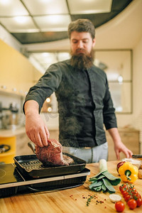 大胡子厨师在厨房的平底锅里煮肉。男人准备煮猪肉在桌上电炉