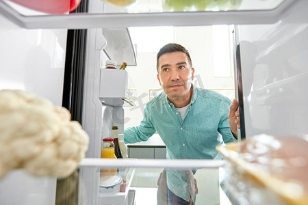 饮食与饮食理念--微笑的中年男子在厨房的冰箱里寻找食物。一名男子在厨房的冰箱里寻找食物