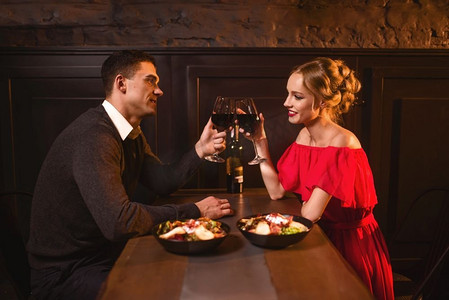 美丽的情侣在餐厅用红酒举起酒杯，浪漫的约会。优雅的女人在红色礼服和她的男人，周年庆祝