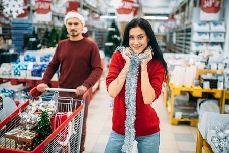 一对年轻夫妇推着购物车在超市的节日装饰部，家庭传统。12月份购买新年或圣诞商品