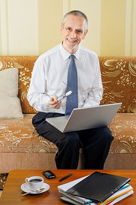 一个英俊的老商人，在家摆弄电脑，手里拿着眼镜，准备喝一杯热咖啡