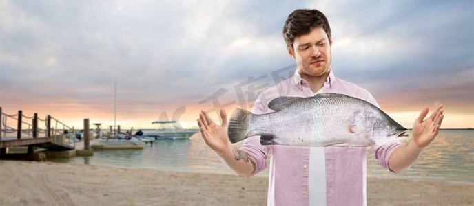 钓鱼和人概念—不愉快的年轻人显示大小的鱼在海滩背景。一名不满的年轻男子在海滩上展示鱼的大小