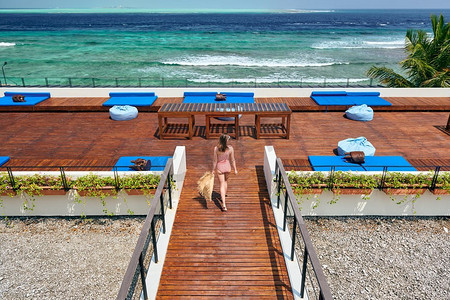 连体比基尼摄影照片_穿着红色条纹连体式泳衣的女子走在热带海滩露台甲板上。在马尔代夫度暑假。