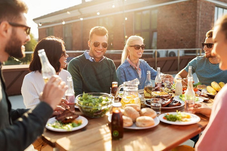 休闲与人的概念-快乐的朋友在天台的烧烤聚会上吃喝。朋友在屋顶上吃晚餐或参加烧烤派对