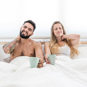 年轻夫妇坐在床上与杯咖啡遭受颈部疼痛