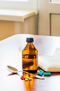 药片摄影照片_靠近窗户的桌子上有一个咳嗽糖浆瓶和一个勺子和一些药片