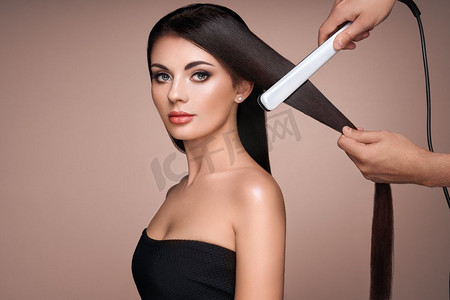 平坦摄影照片_理发师用熨斗拉直长的深色头发。美丽的女人与长的直头发。光滑发型