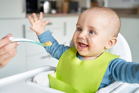 微笑的8个月大的男婴在家里在高脚椅被喂固体食物由母亲用勺子    