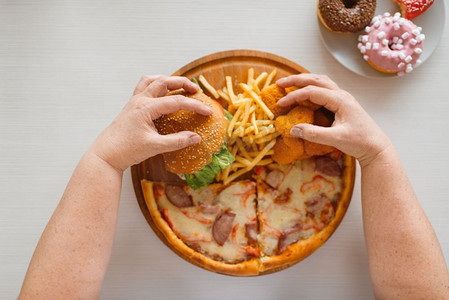 胖女人的手反对高热量食物。超重的女性人在桌子上与垃圾晚餐，肥胖问题，不健康的营养