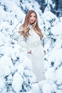 冬日少女摄影照片_冬日森林里的时尚少女。年轻优雅的模特，穿着时髦的白大衣。留着长长光滑头发的金发女孩