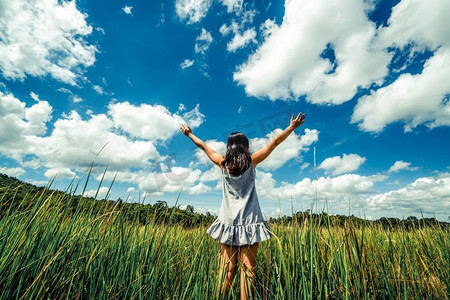 年轻美丽的女人在绿草草甸伸展手臂到蓝色晴朗的天空在夏天。度假和冥想的概念。